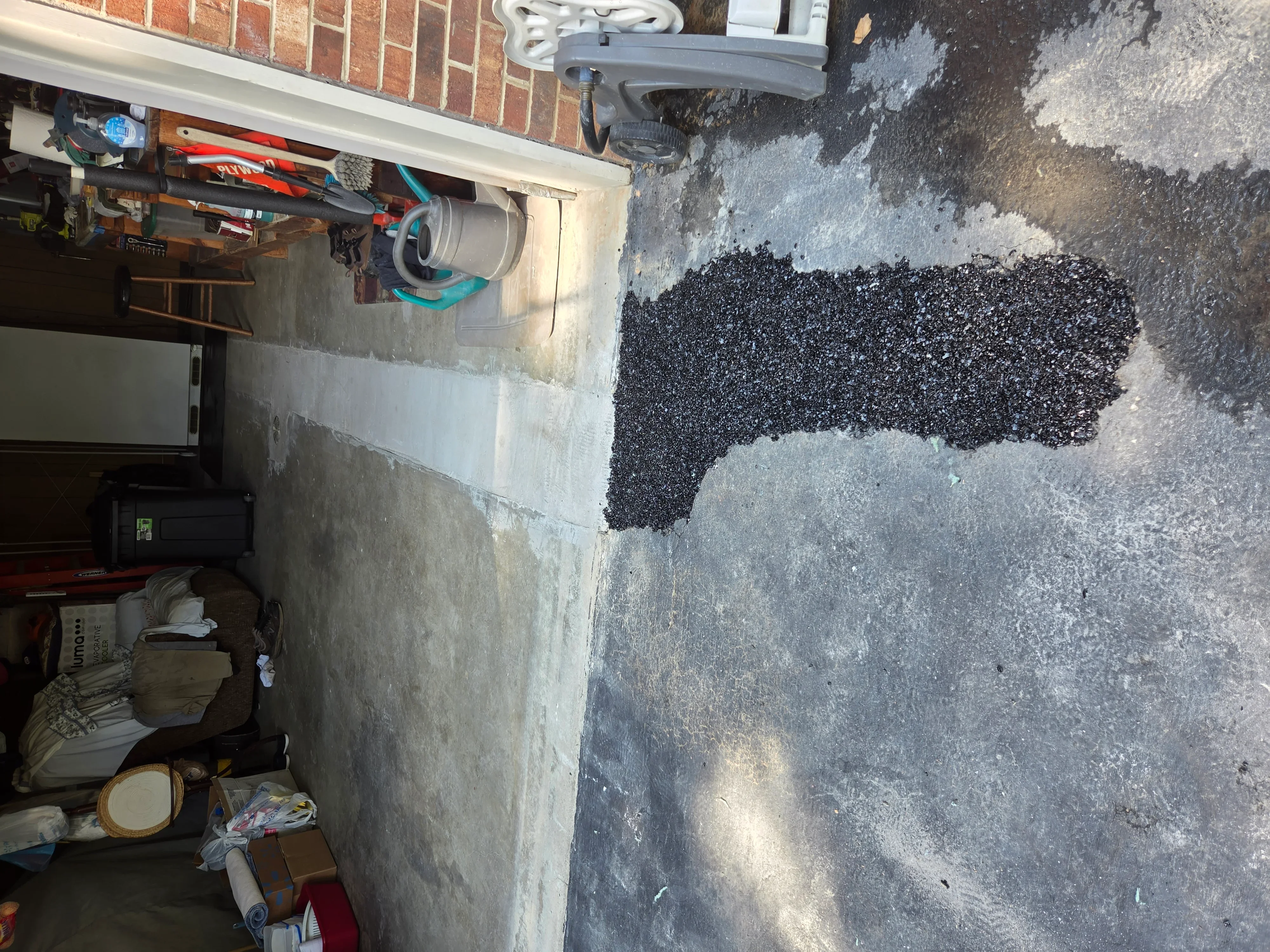 Fresh black asphalt patch applied to worn driveway section next to brick foundation